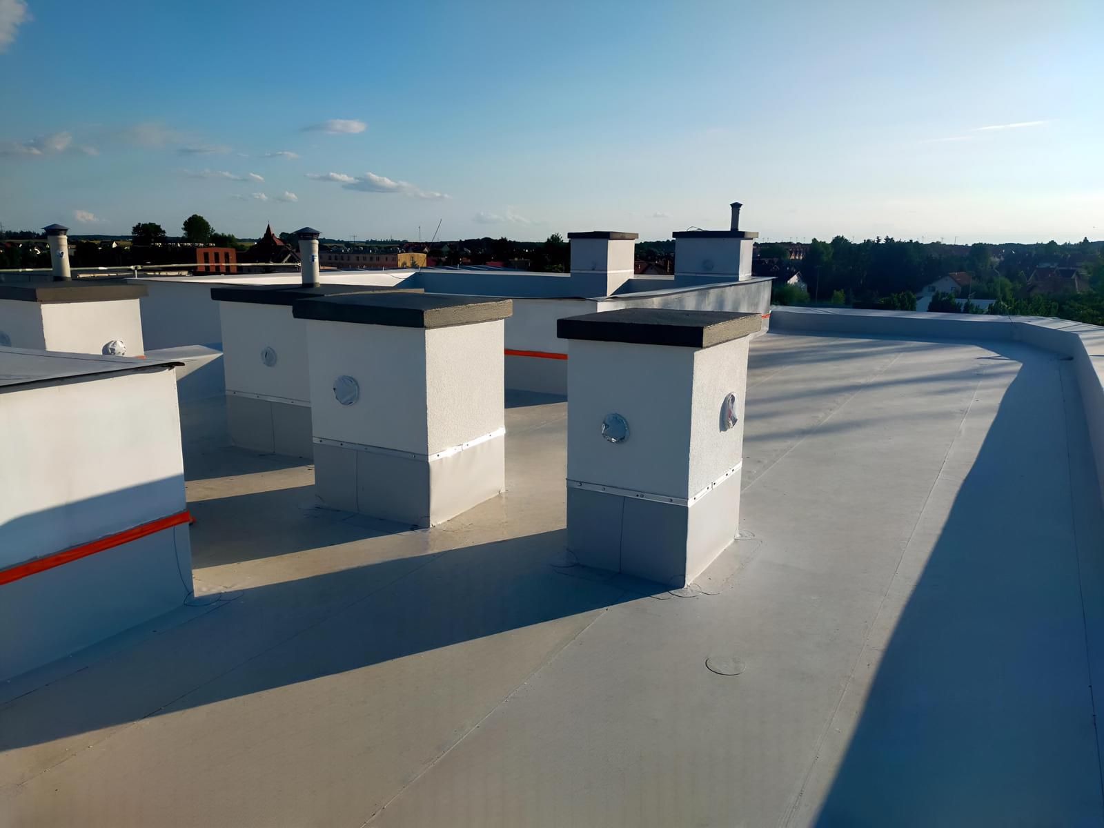Biały, płaski dach z kilkoma kominami wentylacyjnymi o kwadratowej podstawie i szarym wykończeniu, widok na zabudowania i drzewa w tle, jasne niebo.