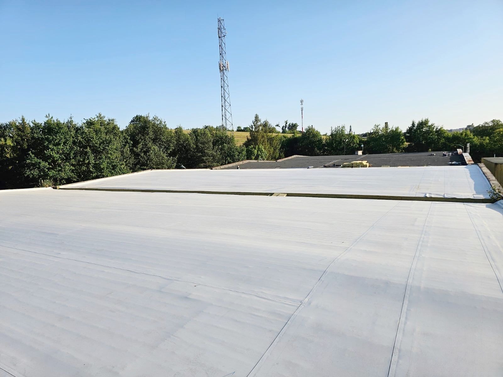 Biała membrana dachowa na płaskim dachu budynku przemysłowego, widok z góry, z zielenią i wieżą telekomunikacyjną w tle.