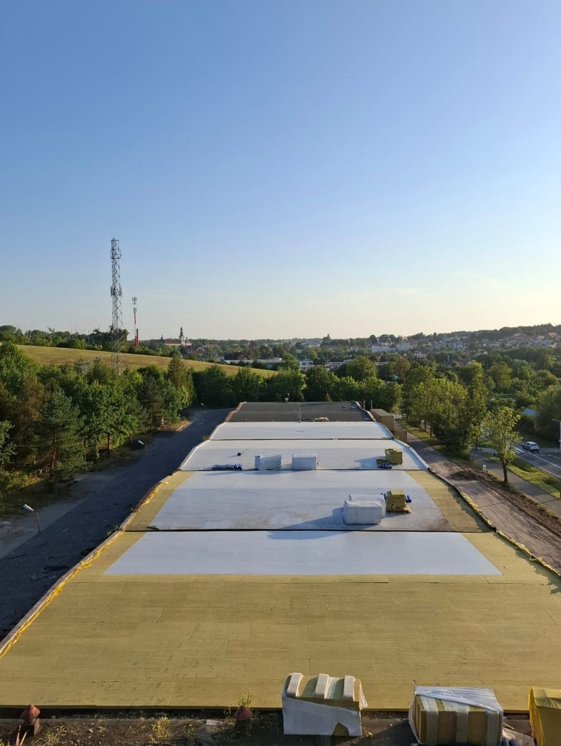 Widok z góry na rozległy dach budynku w trakcie prac izolacyjnych, z widoczną warstwą żółtej wełny mineralnej i białej membrany, na tle zielonego krajobrazu miejskiego i błękitnego nieba.