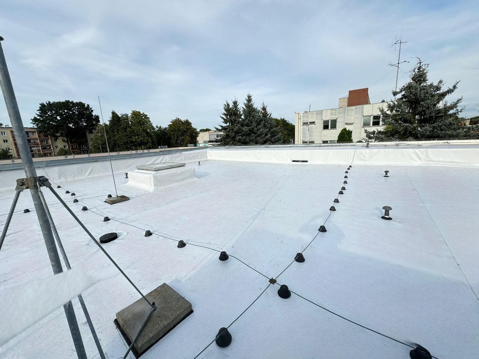 Biały, płaski dach pokryty membraną, z widocznymi elementami mocującymi i zabezpieczeniami, na tle budynków i drzew.