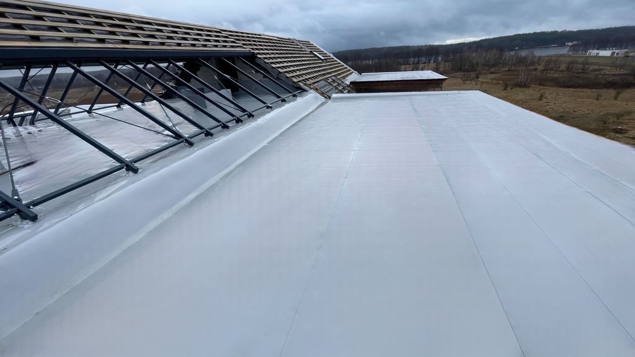 Nowe, białe pokrycie płaskiego dachu z widoczną konstrukcją pod przyszłe przeszklenie, w tle krajobraz z drzewami i jeziorem.