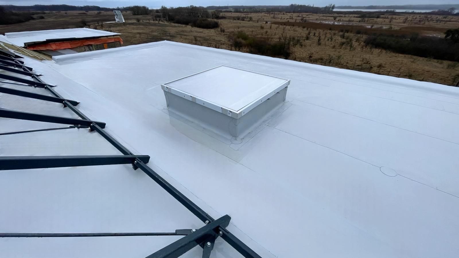 Biały, płaski dach z świetlikiem kwadratowym i elementami konstrukcji w tle, widok z góry.