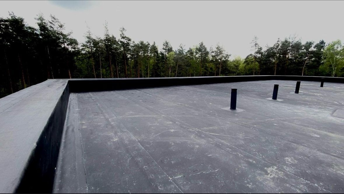 Płaski dach pokryty czarną papą z widocznymi kominkami wentylacyjnymi, w tle zalesiony teren.