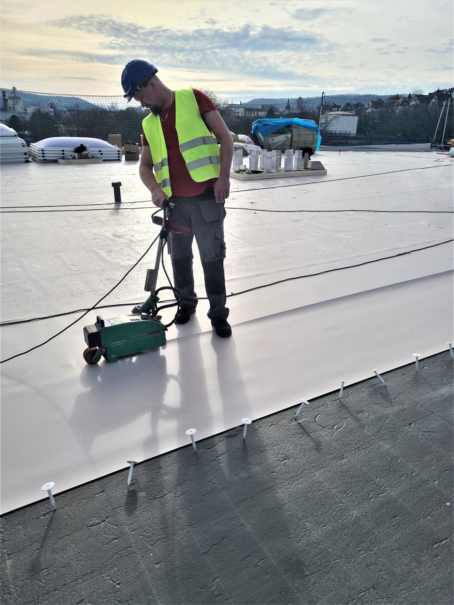 Dekarz w kasku i kamizelce odblaskowej zgrzewa membranę dachową na płaskim dachu za pomocą zgrzewarki VARIMAT V2, widoczne mocowania mechaniczne membrany.