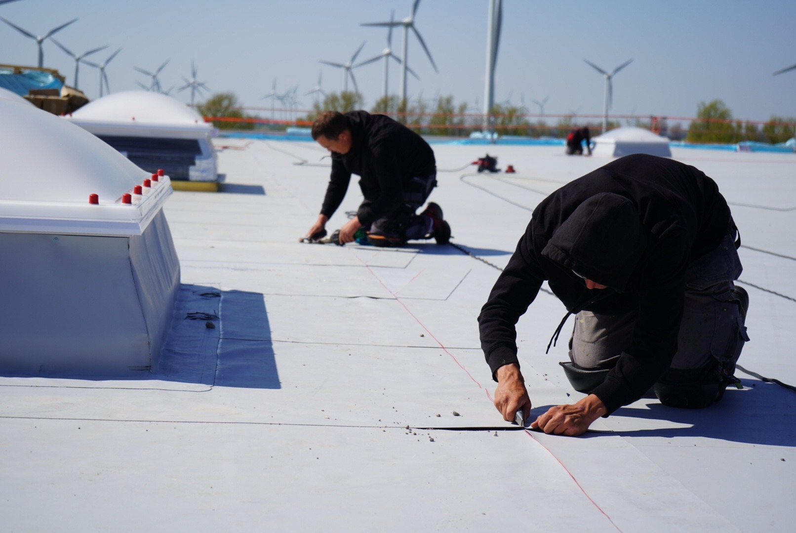 Pracownicy na dachu z białej membrany, przycinają materiał nożem, w tle turbiny wiatrowe.