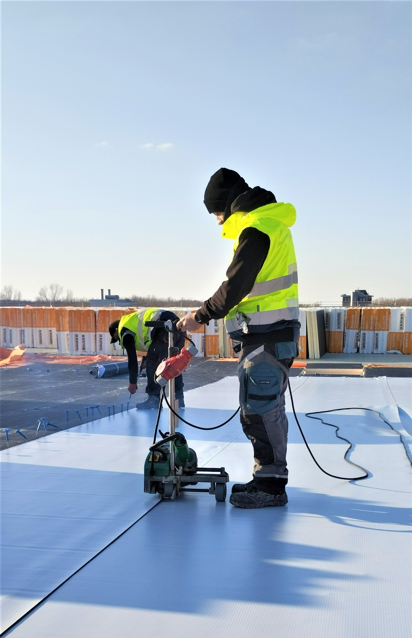 Dekarze zgrzewają membranę dachową na płaskim dachu za pomocą zgrzewarki automatycznej, w tle izolacja termiczna BauderPIR.