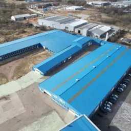 Zdjęcie z drona przedstawia kompleks budynków przemysłowych z niebieskimi dachami, z widocznymi oknami i drzwiami, otoczonych parkingiem i terenem zielonym.