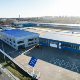 Widok z lotu ptaka na budynki fabryki okien i drzwi, z niebieskim dachem i logo 'illuminator', tablica informacyjna z napisem 'Fabryka okien i drzwi PVC Alu' na pierwszym planie, samochody i pieszy...