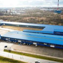 Widok z lotu ptaka na duży, nowoczesny zakład produkcyjny z niebieskim dachem, otoczony zielenią i parkingiem z samochodami, na tle miejskiego krajobrazu z kominem fabrycznym.