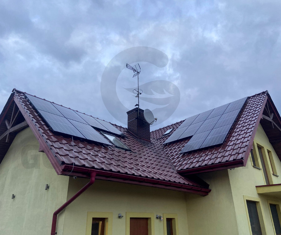 Panele słoneczne zamontowane na dachu domu jednorodzinnego z czerwoną dachówką, widoczna antena satelitarna i telewizyjna, pochmurne niebo w tle.
