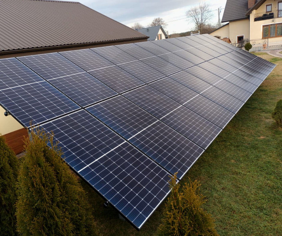 Instalacja fotowoltaiczna naziemna, panele ustawione pod kątem na trawniku przed domem jednorodzinnym z brązowym dachem, widoczne małe tuje.