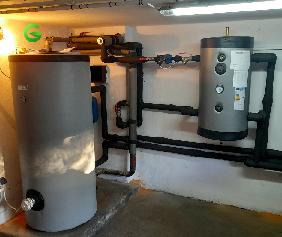 Dwa szare zbiorniki akumulacyjne ciepła połączone rurami w piwnicy, widoczny manometr i etykieta energetyczna na mniejszym zbiorniku.