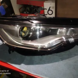 Reflektor samochodowy z soczewką, widoczny na tle roboczym, z pudełkami i narzędziami w tle.