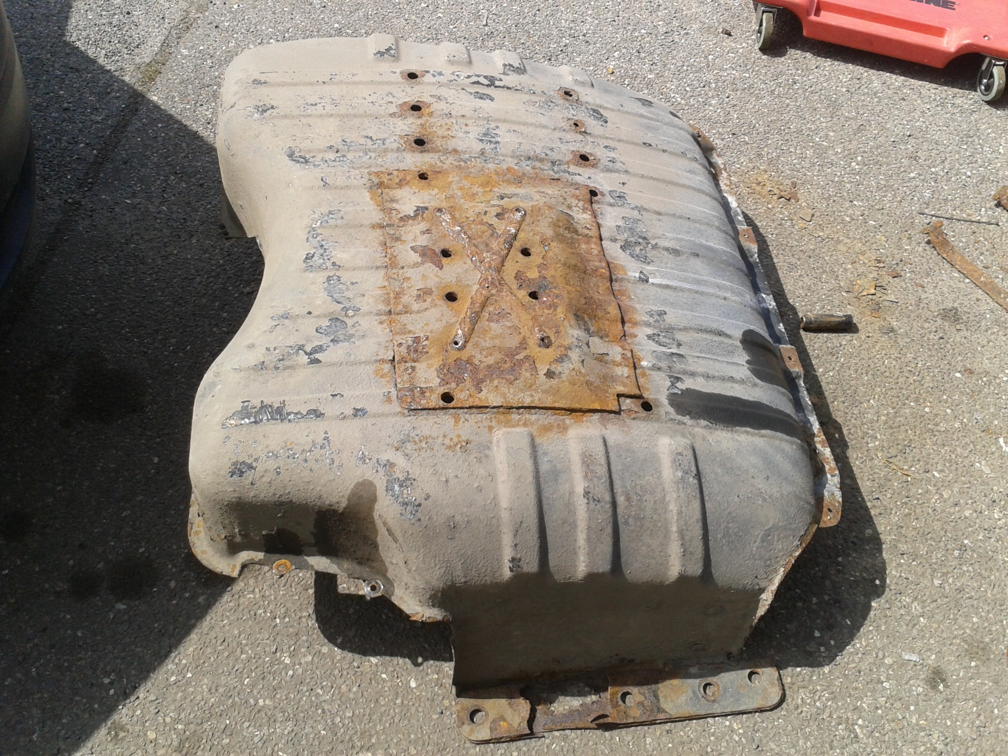 Zdemontowany, skorodowany zbiornik paliwa samochodowego z widocznymi śladami rdzy i napraw blacharskich, leżący na betonowym podłożu w warsztacie.