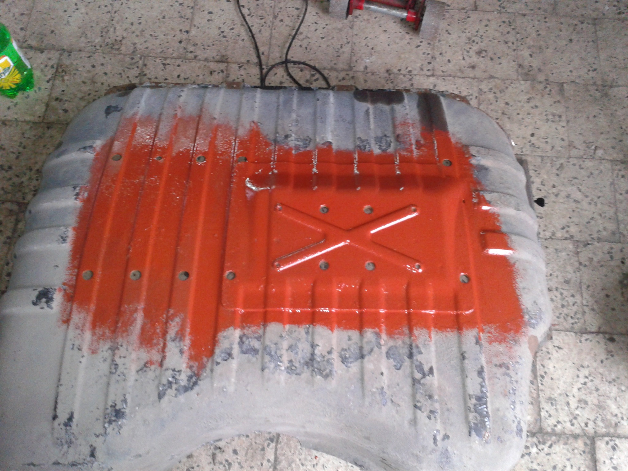 Częściowo pomalowany na pomarańczowo zbiornik paliwa z widocznymi śladami korozji, leżący na podłodze z płytek.