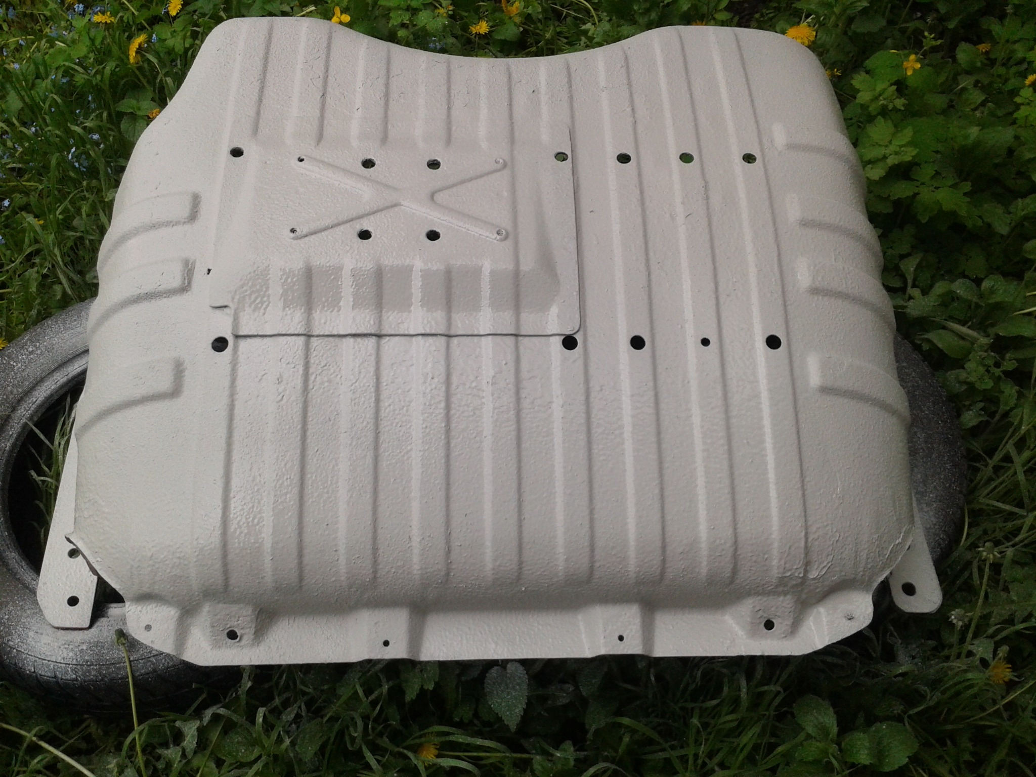 Metalowa osłona podwozia samochodu po odnowieniu, pomalowana na jasnoszary kolor, leży na trawie obok opony.