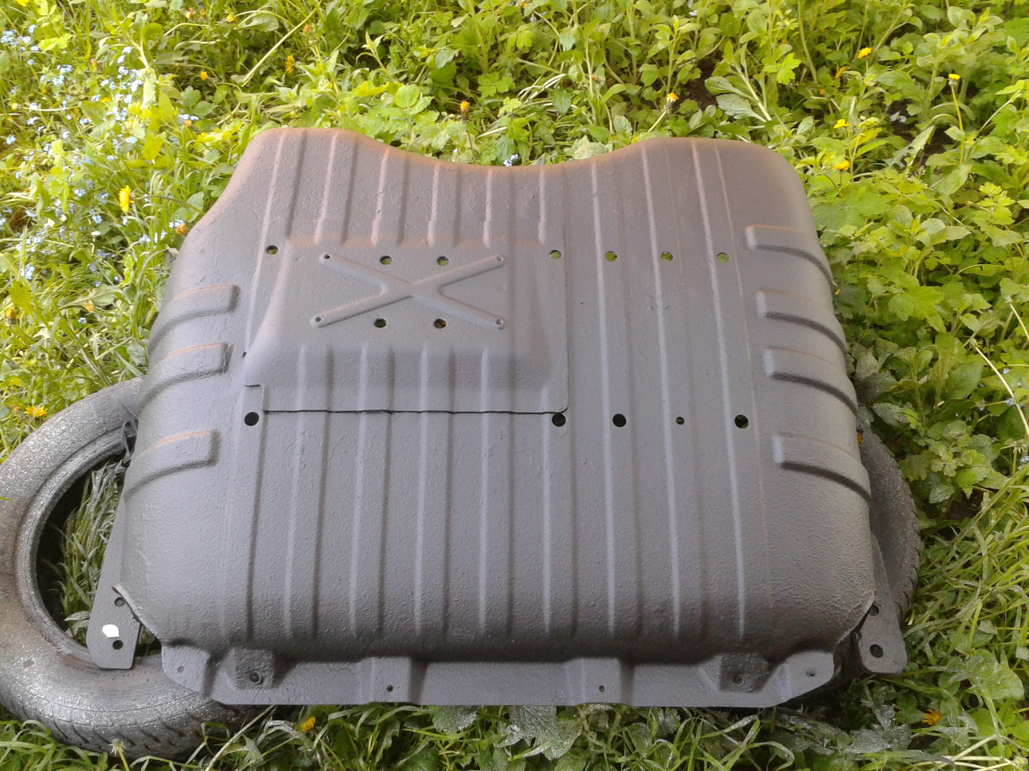 Metalowa osłona podwozia po piaskowaniu i malowaniu, leżąca na trawie obok opony samochodowej. Widoczne ślady chropowatej faktury po malowaniu.