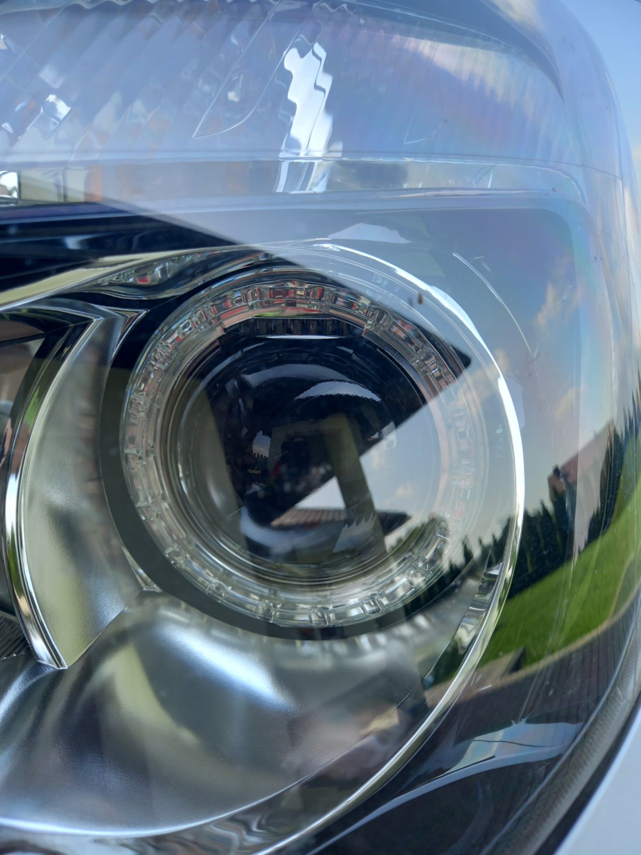 Szczegółowe zbliżenie na reflektor samochodowy z soczewką projektorową, pokazujący odbicie otoczenia i precyzyjne detale konstrukcyjne.