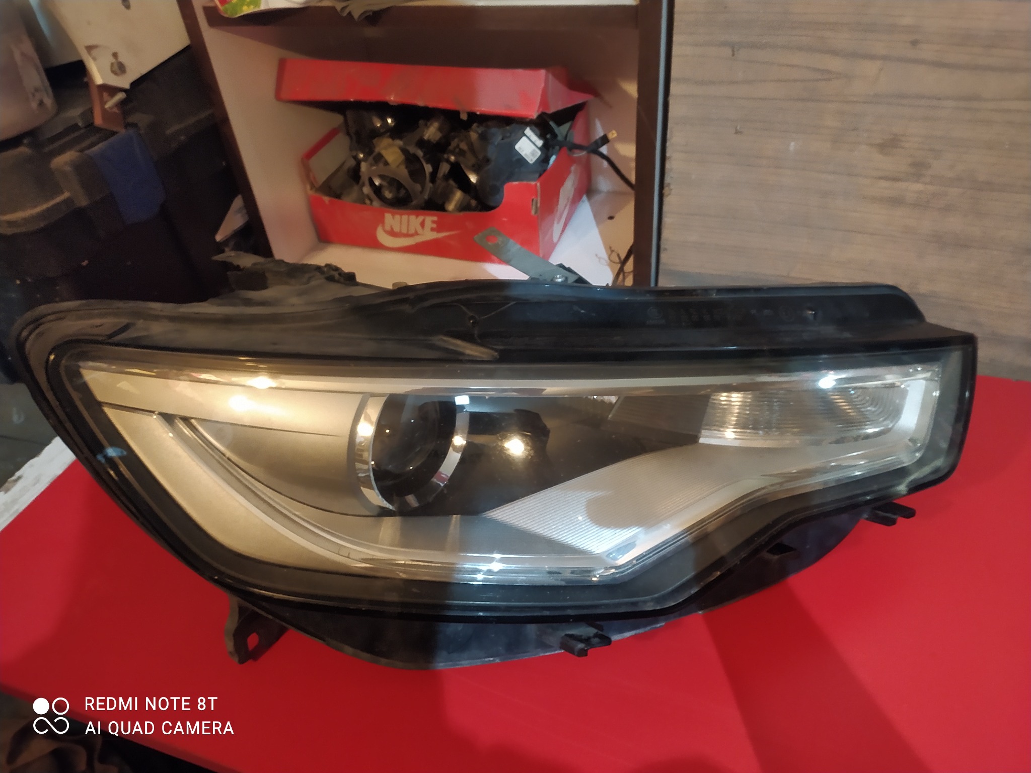 Używana lampa samochodowa, widoczne ślady użytkowania, tło z elementami warsztatowymi, pudełko po butach Nike z częściami samochodowymi w tle.