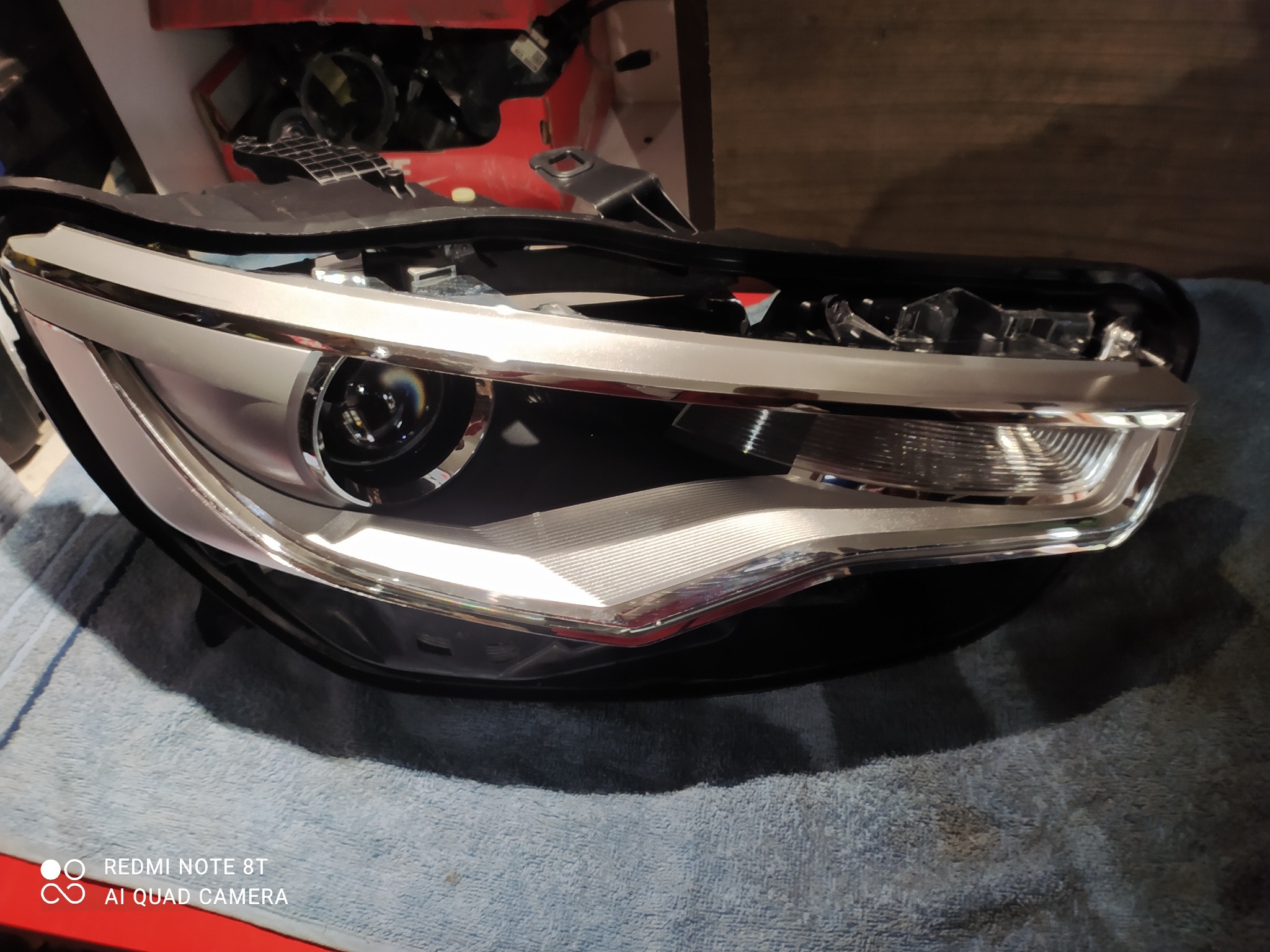 Reflektor samochodowy z chromowanymi elementami, leżący na szarej tkaninie, widoczne delikatne zabrudzenia i niedoskonałości.