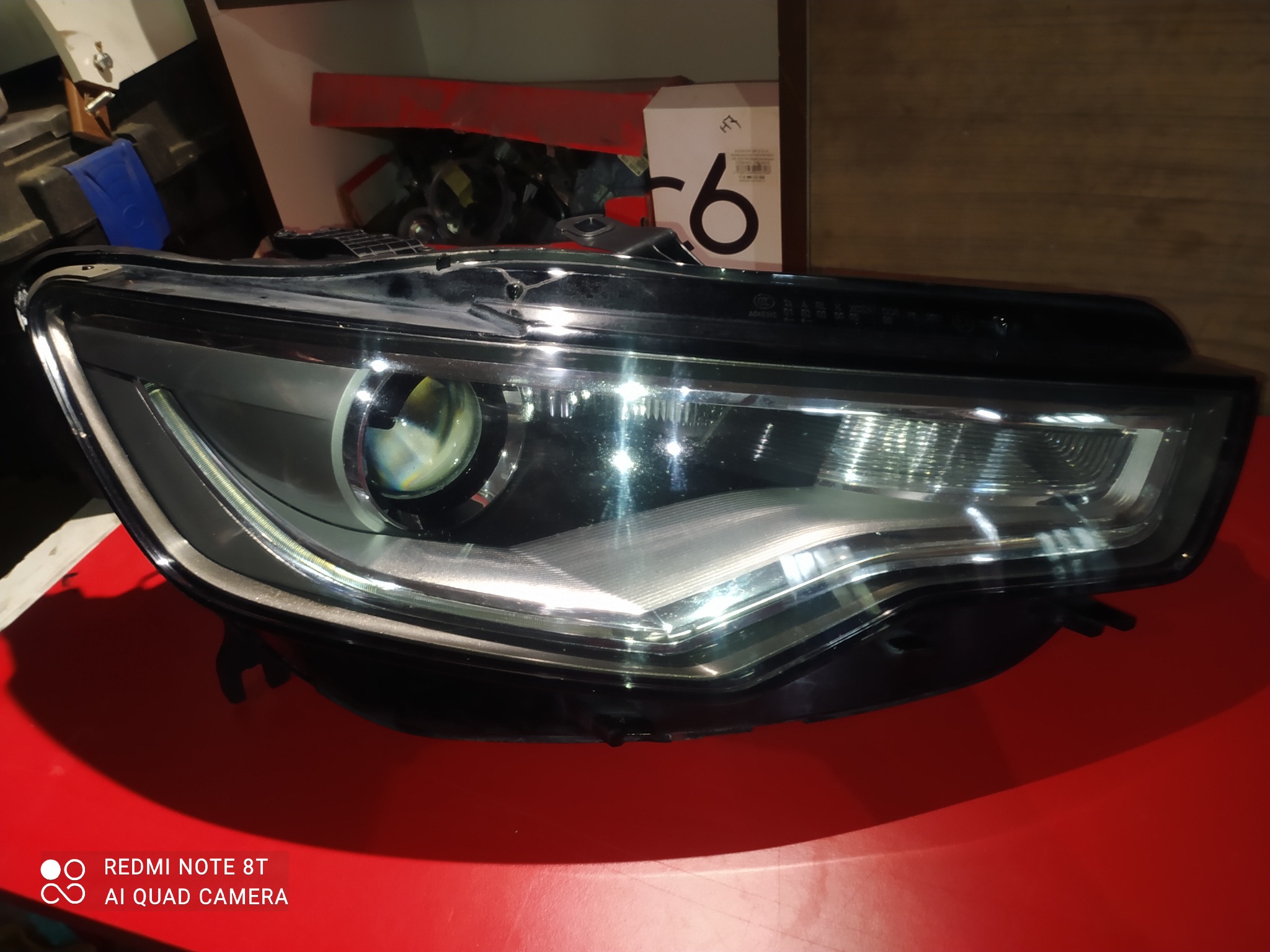 Reflektor samochodowy na czerwonym tle, widoczne detale konstrukcyjne i odbicia światła, obok pudełko z cyfrą 6.