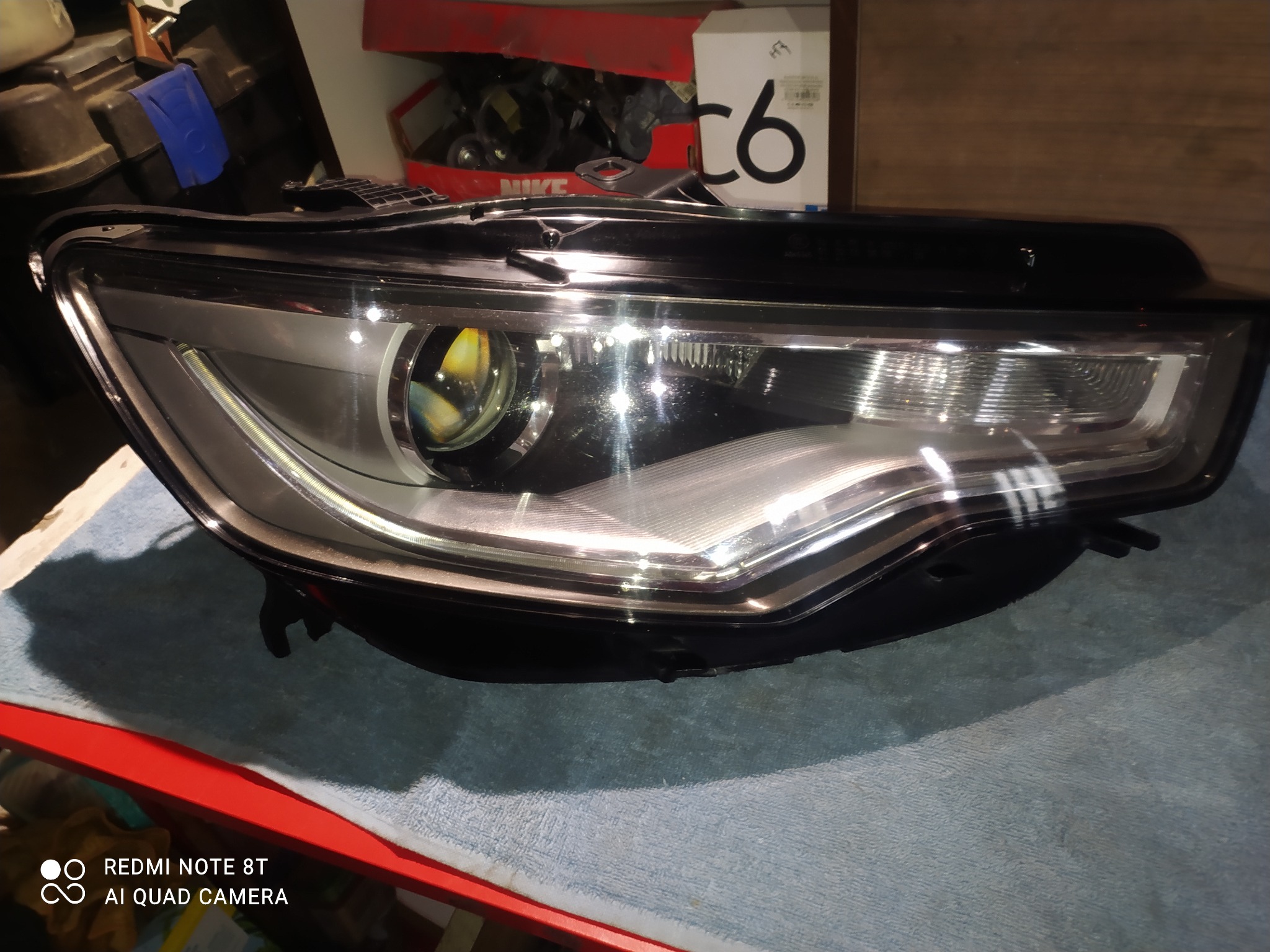 Reflektor samochodowy z soczewką, widoczny na tle roboczym, z pudełkami i narzędziami w tle.