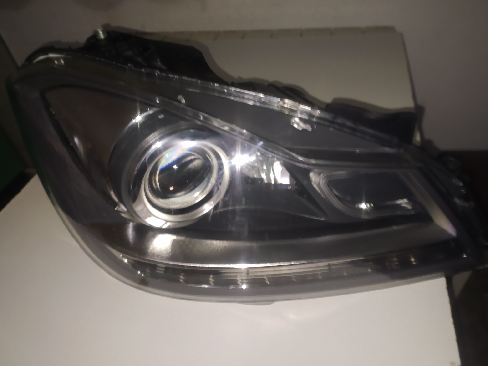 Reflektor samochodowy z widocznymi soczewkami i elementami mocującymi, leżący na białej powierzchni, oświetlony z góry.