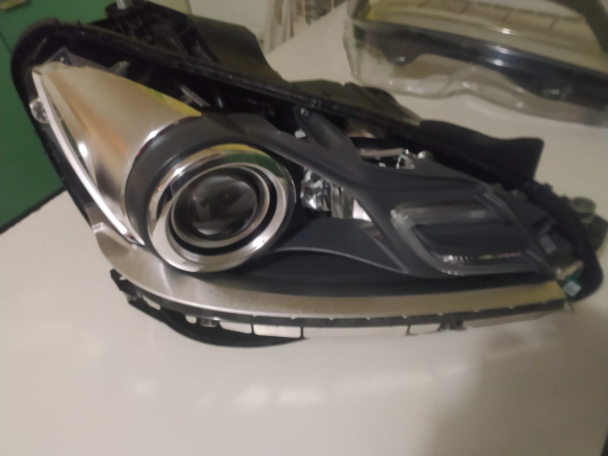 Reflektor samochodowy z widocznymi elementami chromowanymi i czarną obudową, leżący na jasnym tle.