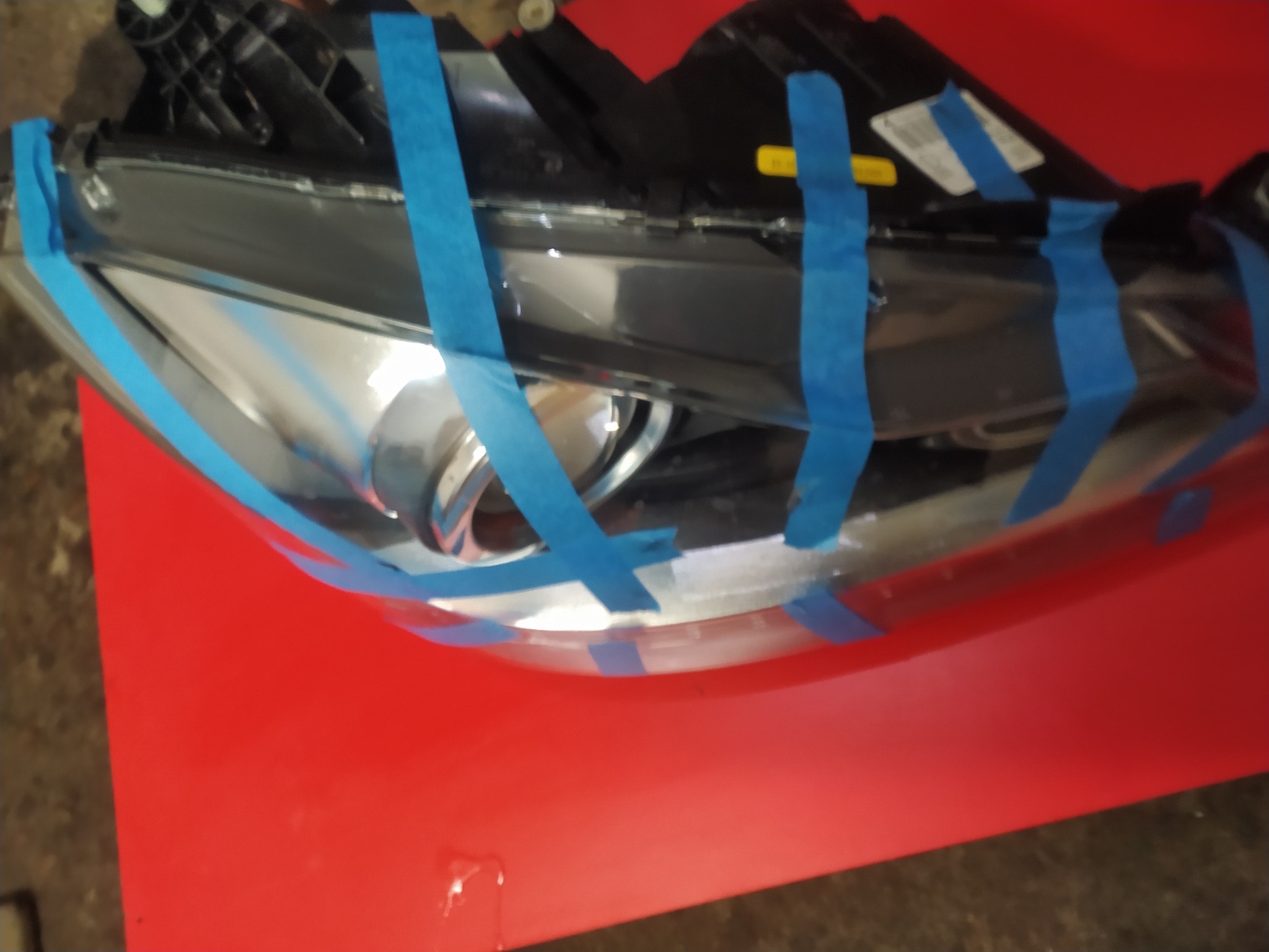 Reflektor samochodowy zabezpieczony niebieską taśmą malarską, przygotowany do renowacji na czerwonym podłożu.