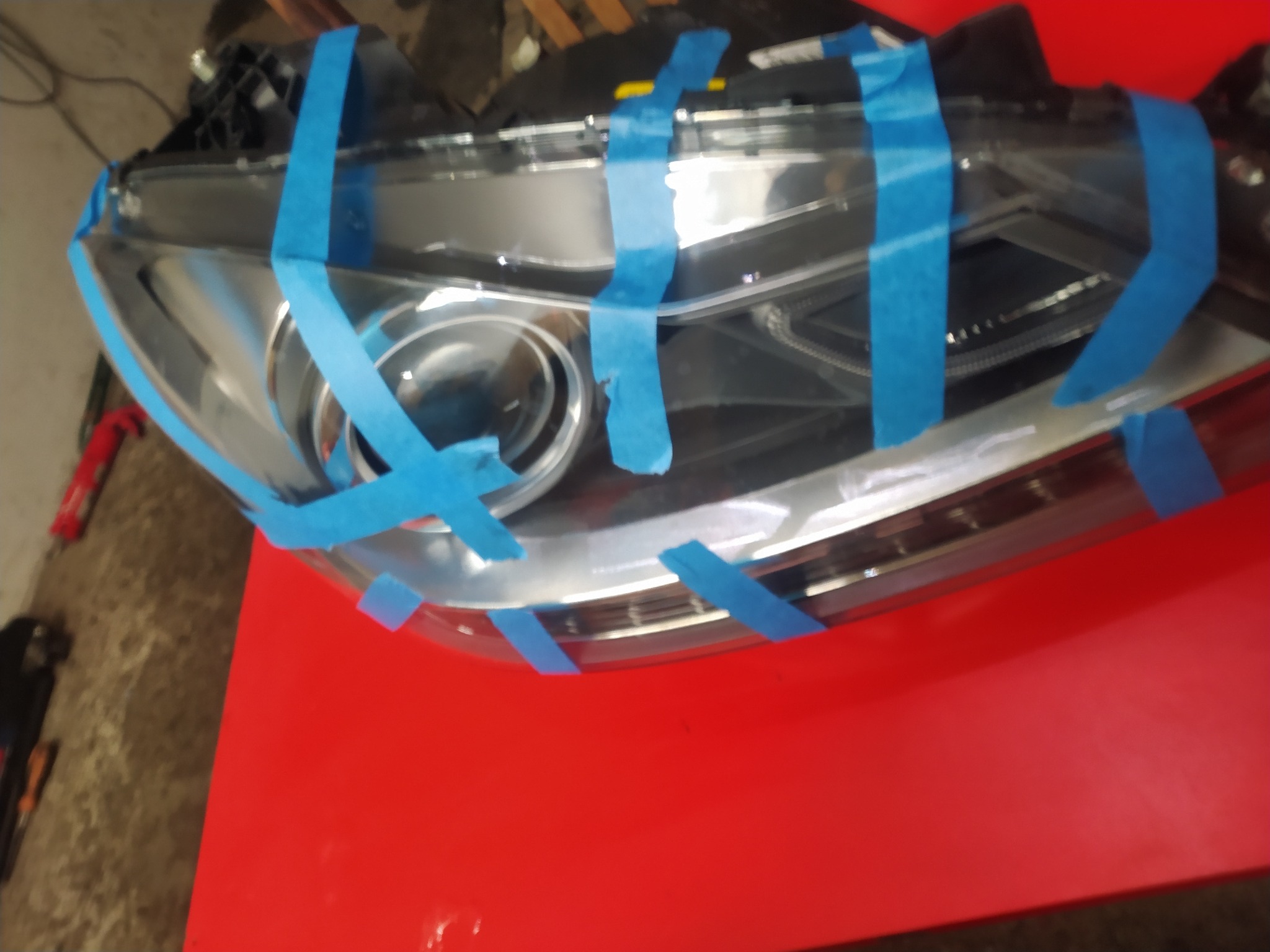 Reflektor samochodowy zabezpieczony niebieską taśmą malarską przed obróbką, leżący na czerwonym stole warsztatowym.