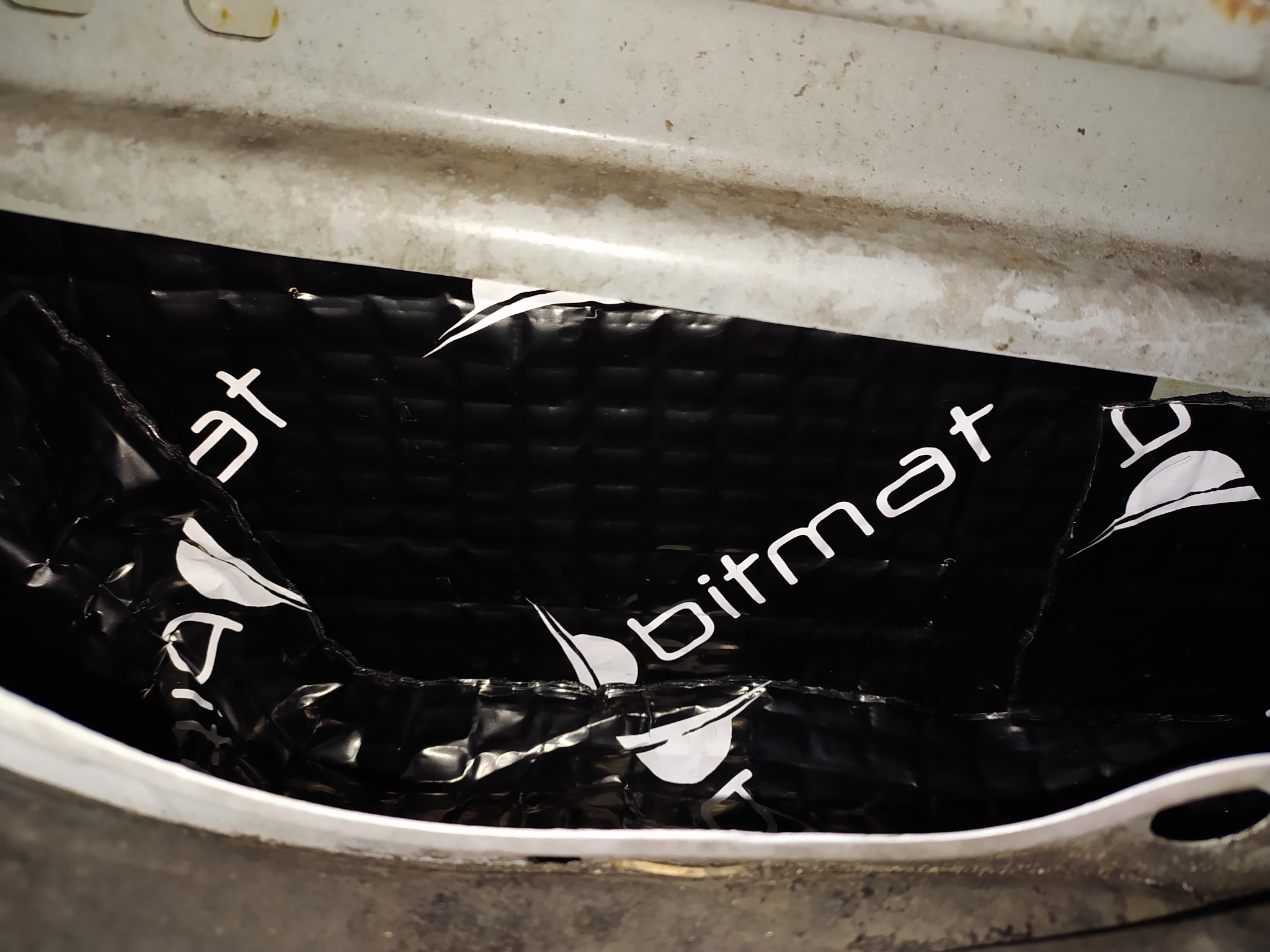 Aplikacja maty wygłuszającej Bitmat wewnątrz nadkola samochodu, widoczne niedoskonałości montażu i zabrudzenia na metalowych elementach.