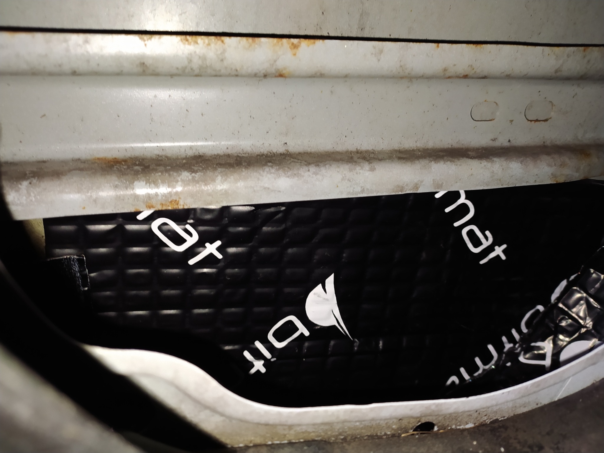 Fragment wnętrza drzwi samochodu z naklejoną czarną matą wygłuszającą z białym napisem 'bitmat', widoczne ślady korozji na metalowych elementach.