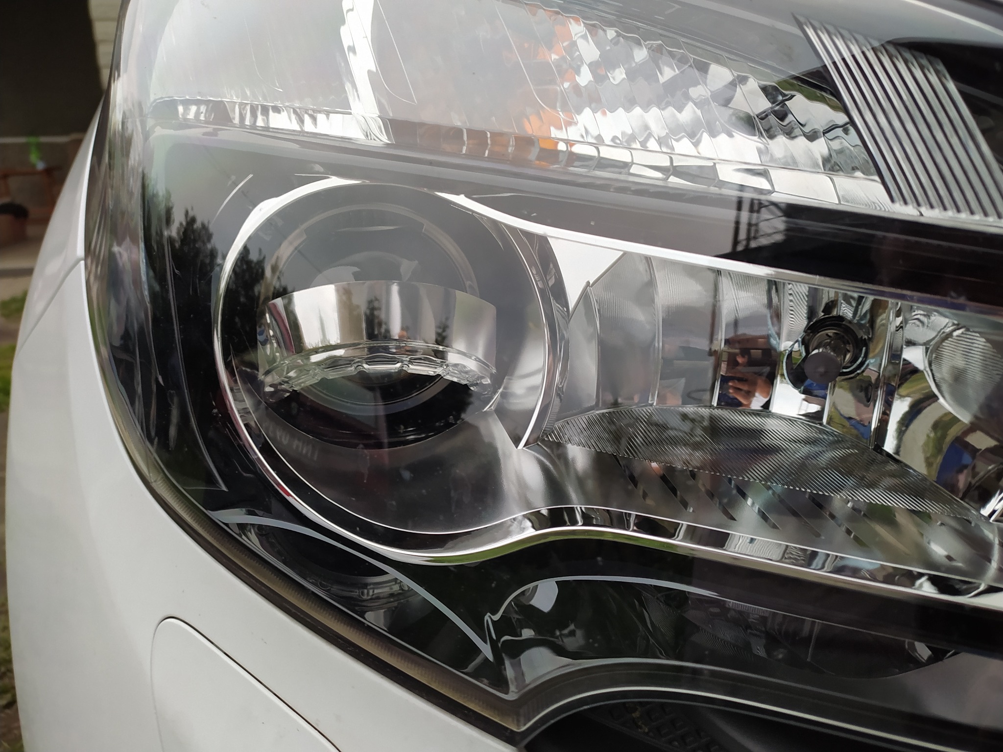 Szczegółowe ujęcie reflektora nowoczesnego białego samochodu, widoczne odbicia otoczenia na gładkiej powierzchni klosza.