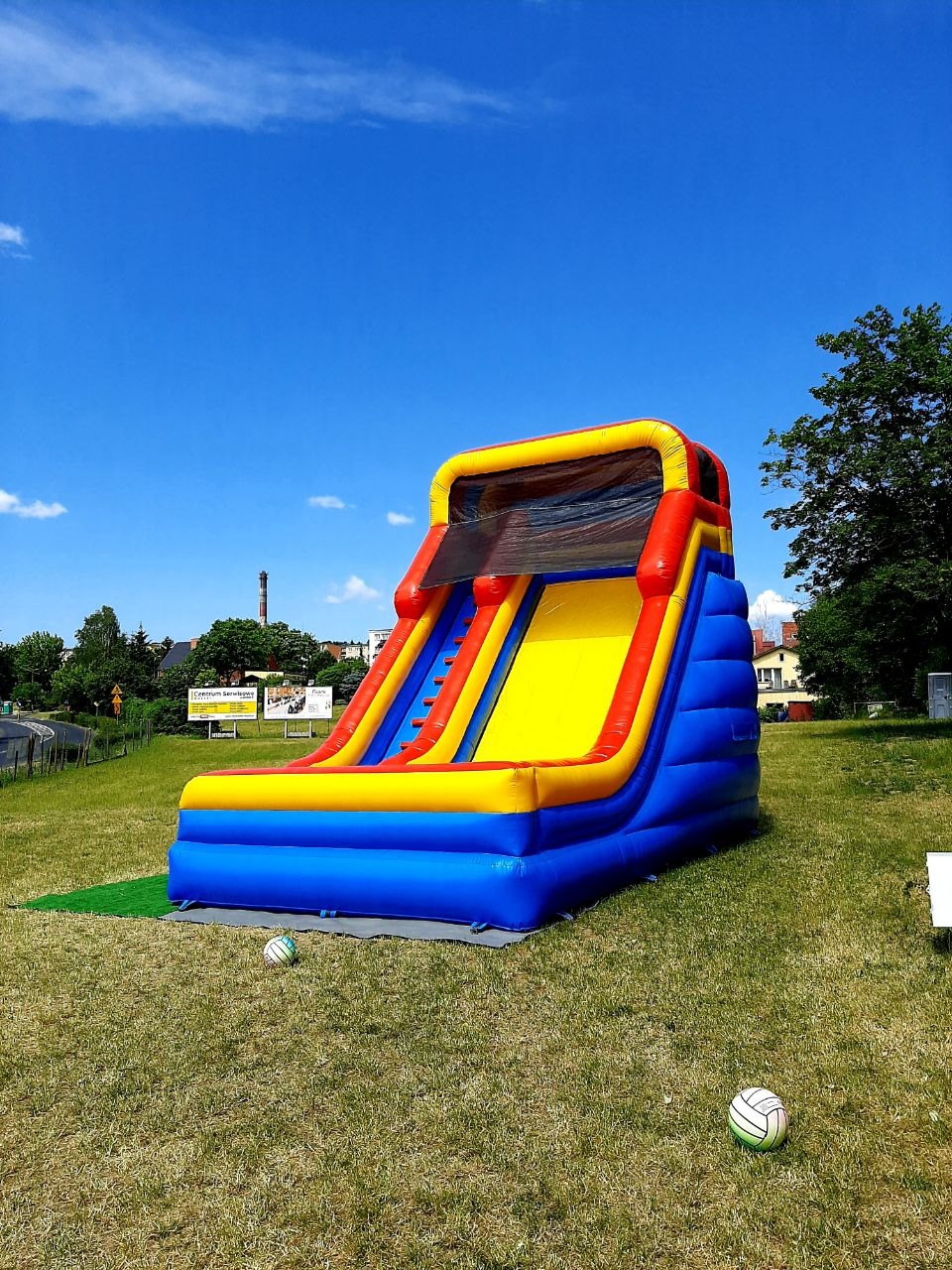 Kolorowa dmuchana zjeżdżalnia z żółtym ślizgiem, czerwonymi bokami i niebieską podstawą, ustawiona na trawie obok dwóch piłek, w tle błękitne niebo i drzewa.