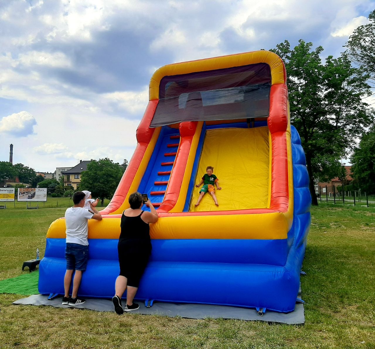 Chłopiec zjeżdża na żółtej zjeżdżalni dmuchanej o konstrukcji niebiesko-żółto-czerwonej; rodzice robią zdjęcia na tle zielonego trawnika i budynków.