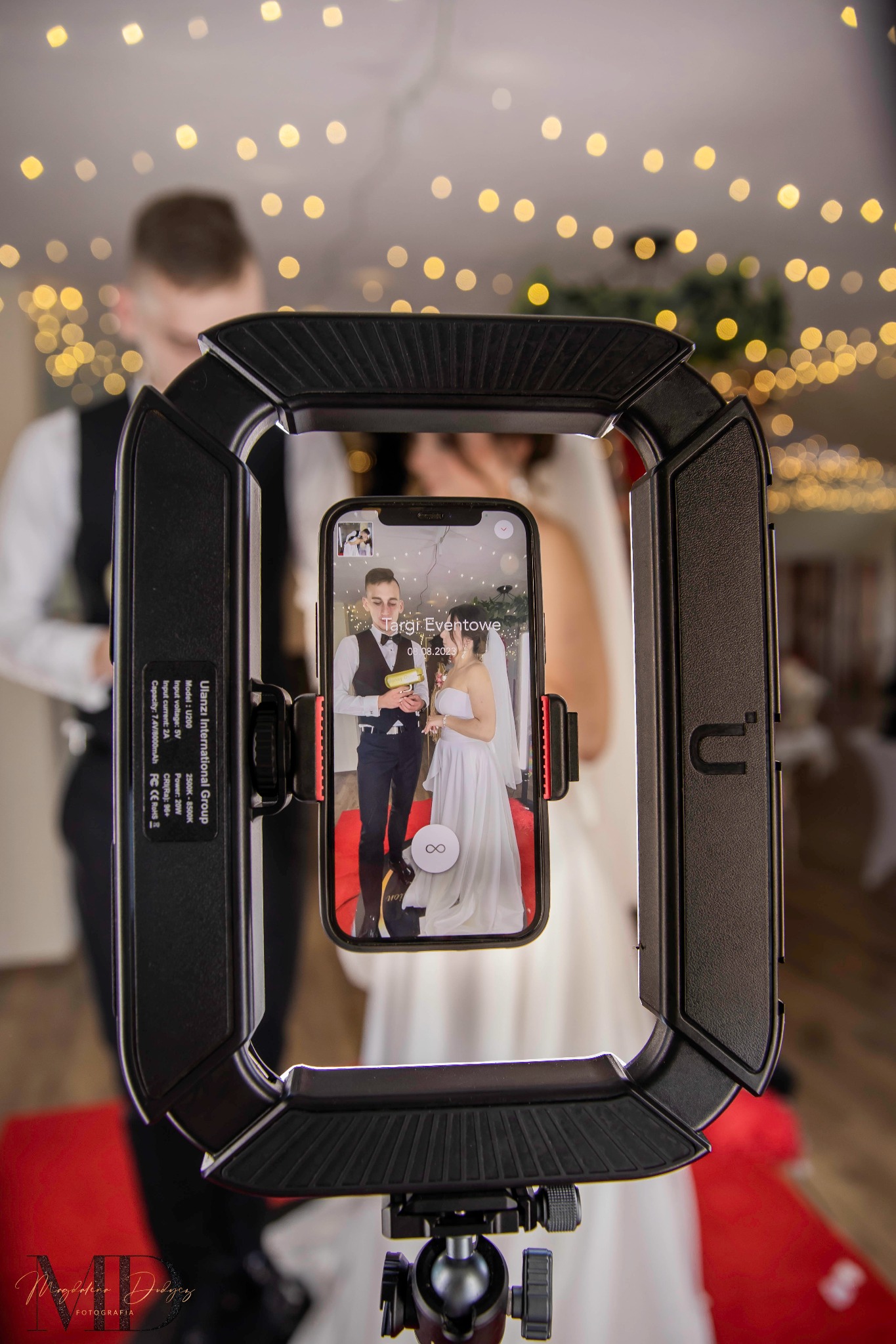 Smartfon w uchwycie oświetleniowym na statywie, wyświetlający nagranie wideo z elegancko ubraną parą młodą w trakcie wznoszenia toastu na tle sali z dekoracją świetlną.