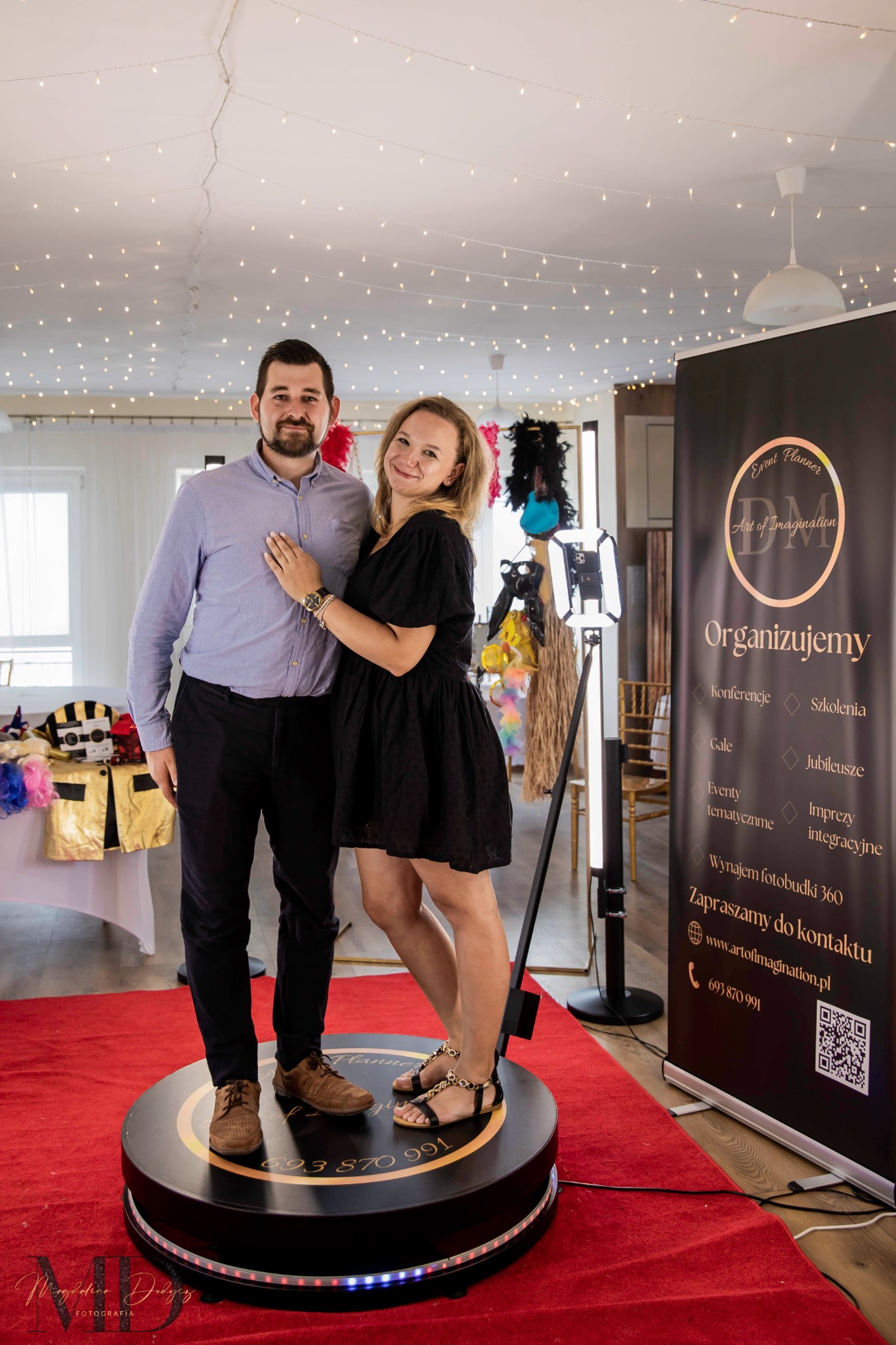 Para pozuje na obrotowej platformie fotobudki 360 z oświetleniem LED, na czerwonym dywanie, w tle rekwizyty i baner reklamowy z ofertą konferencji, szkoleń i imprez integracyjnych.
