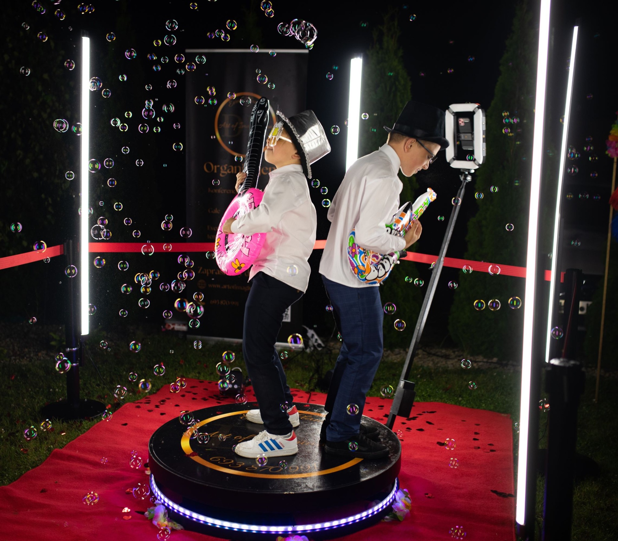Dwaj chłopcy w zabawnych kapeluszach i z dmuchanymi gitarami pozują na obrotowej platformie fotobudki 360, otoczeni bańkami mydlanymi i oświetleni lampami LED.