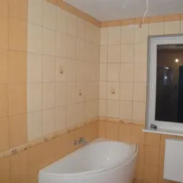 Wykończona łazienka z beżowymi i pomarańczowymi płytkami, białą wanną narożną i grzejnikiem pod oknem. Widoczne niedociągnięcia w postaci wystających kabli i brakujących fug.