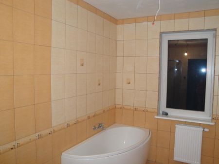 Wykończona łazienka z beżowymi i pomarańczowymi płytkami, białą wanną narożną i grzejnikiem pod oknem. Widoczne niedociągnięcia w postaci wystających kabli i brakujących fug.