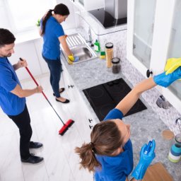 Agencja Leone - Zespół sprzątający w niebieskich uniformach: mężczyzna zamiata podłogę czerwoną szczotką, kobieta myje zlew, a kolejna kobieta czyści szafki kuchenne żółtą szmatką w niebieskich rękawicach.