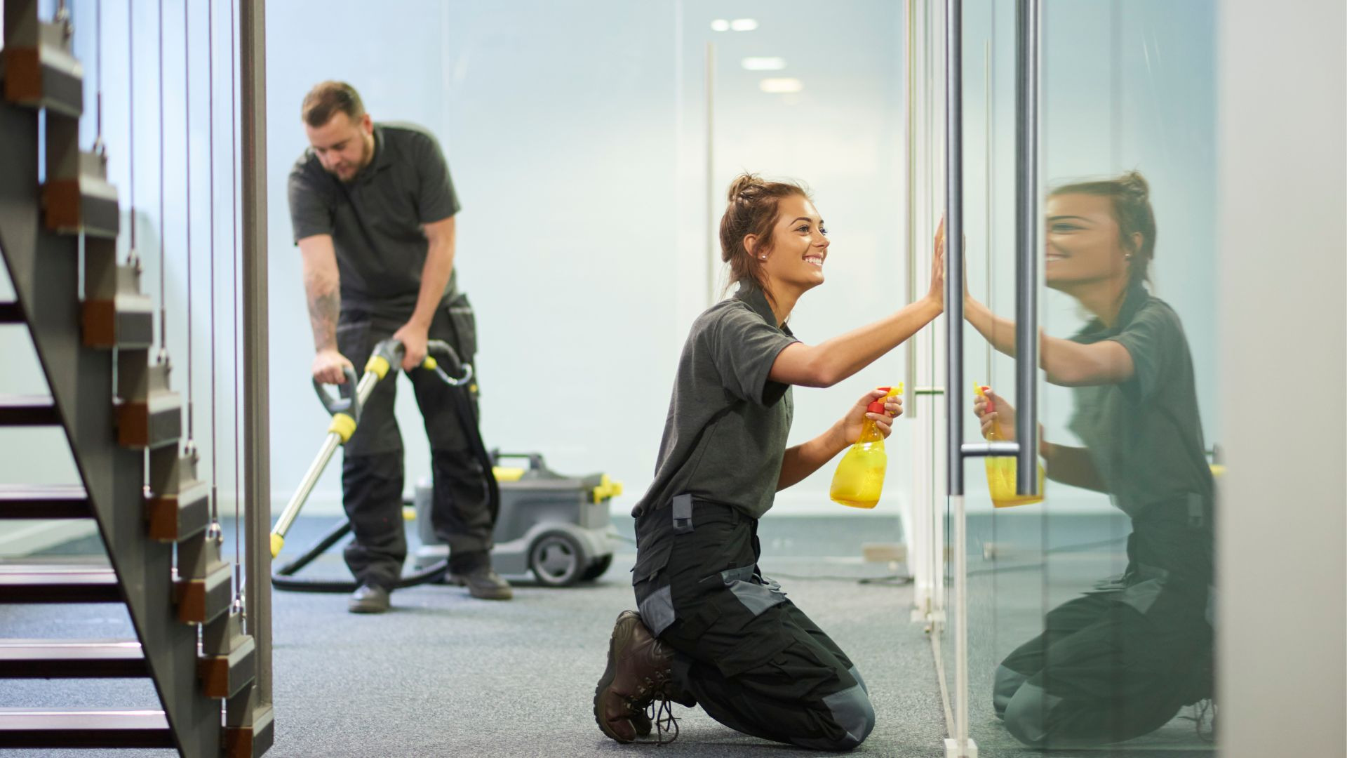Uśmiechnięta kobieta na kolanach czyści szklaną ścianę, w tle mężczyzna odkurza podłogę, widoczny odkurzacz przemysłowy i schody.