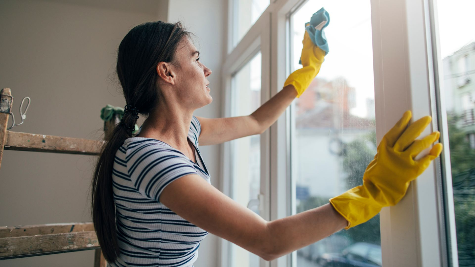 Kobieta w żółtych rękawicach czyści okno szmatką, z boku widoczna drewniana drabina malarska.