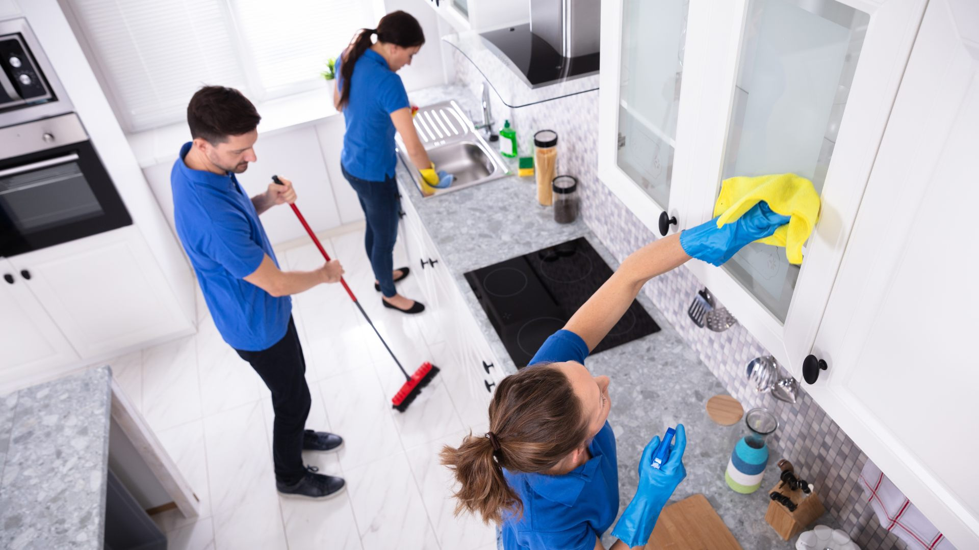 Zespół sprzątający w niebieskich uniformach: mężczyzna zamiata podłogę czerwoną szczotką, kobieta myje zlew, a kolejna kobieta czyści szafki kuchenne żółtą szmatką w niebieskich rękawicach.