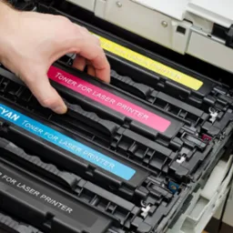 Wymiana kolorowych tonerów w drukarce laserowej; widoczne wkłady z oznaczeniami: czarny, cyan, magenta i żółty, w trakcie instalacji przez dłoń wewnątrz otwartej obudowy drukarki.