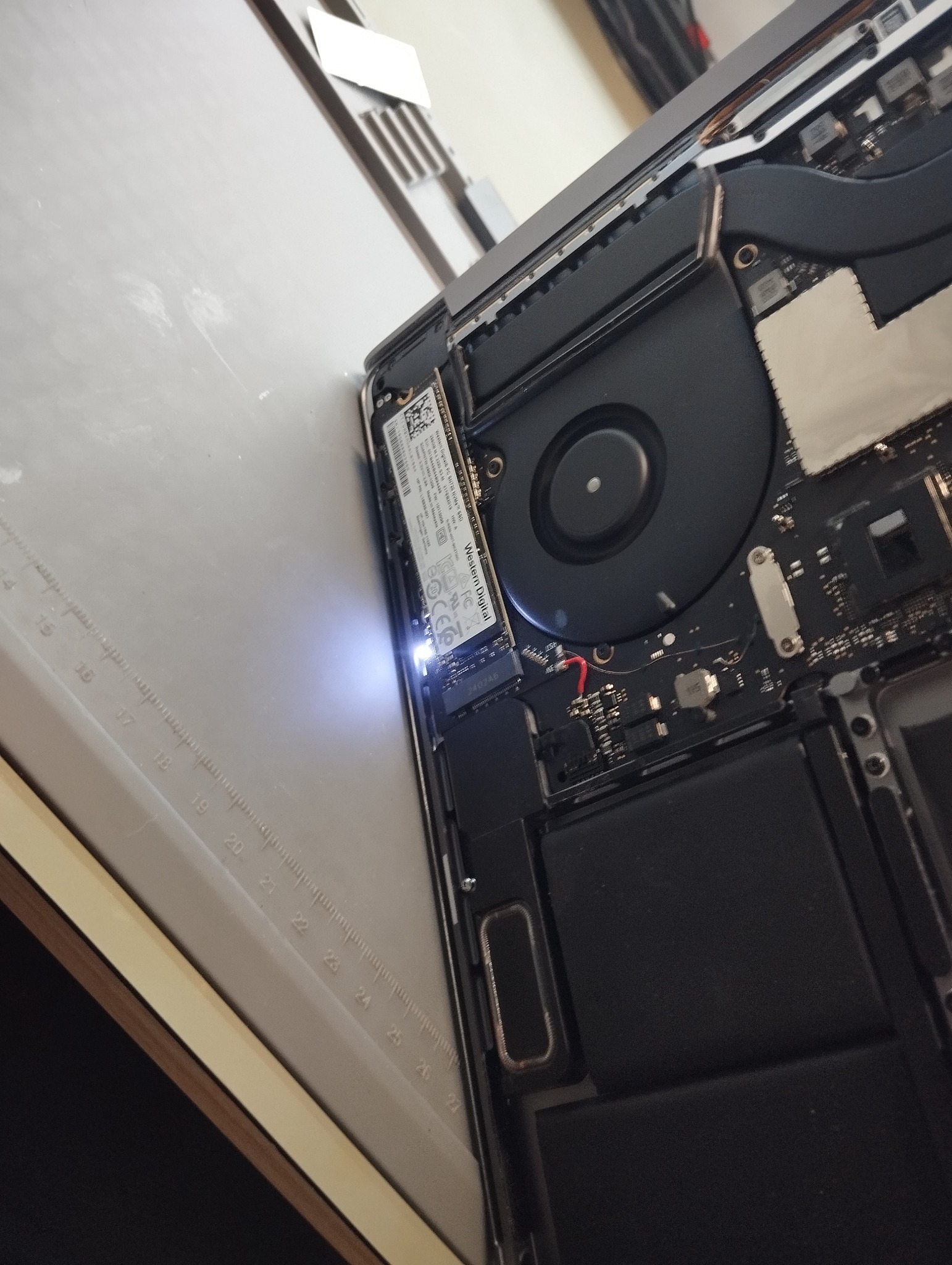 Otwarty laptop Apple z widocznym dyskiem SSD Western Digital, wentylatorem i elementami płyty głównej, oświetlony punktowo. Widok z góry, lekki kąt.