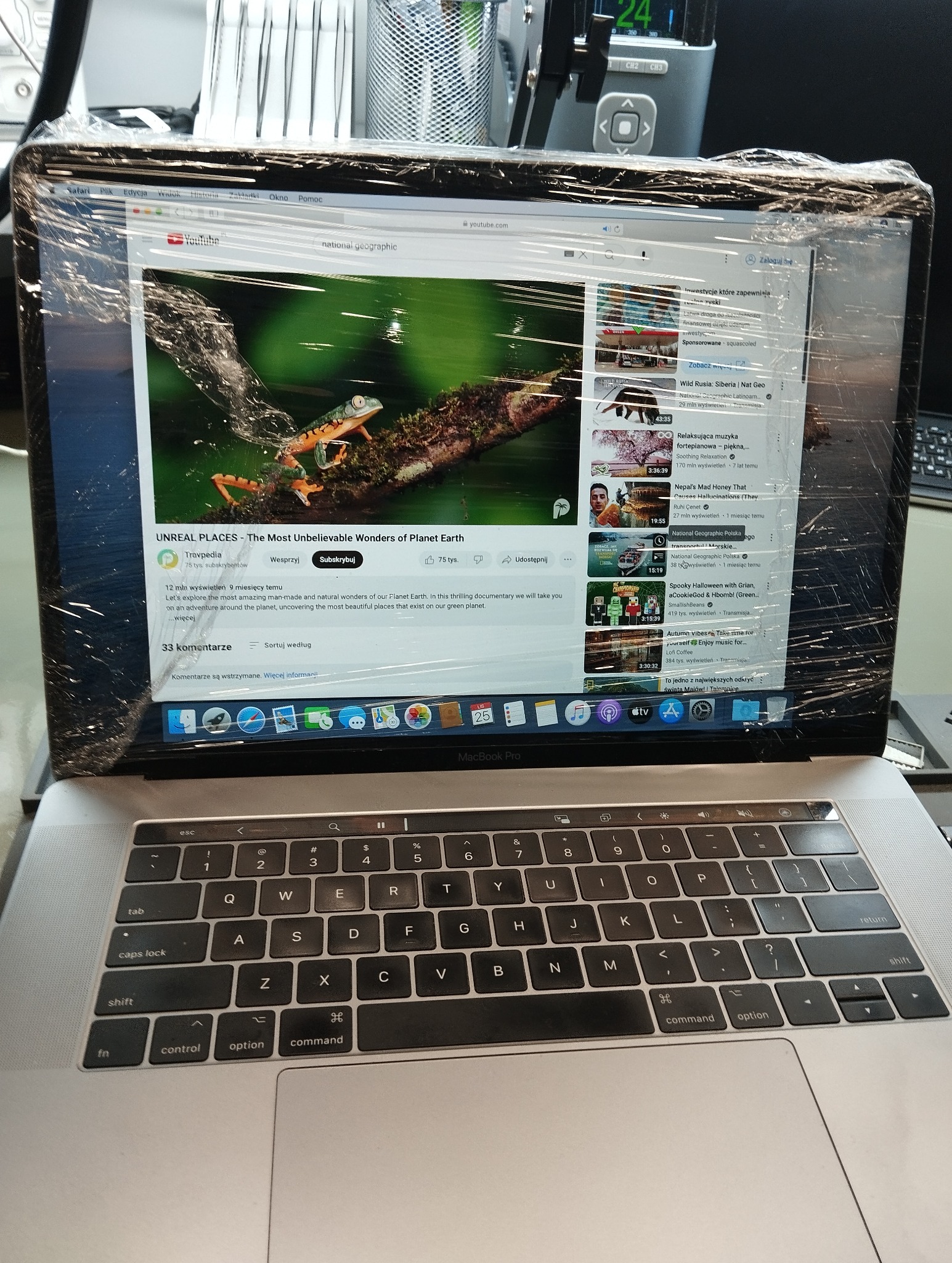 Laptop MacBook Pro z włączonym YouTube, ekran zabezpieczony folią. Widoczny film National Geographic o cudach natury. Klawiatura w tle.