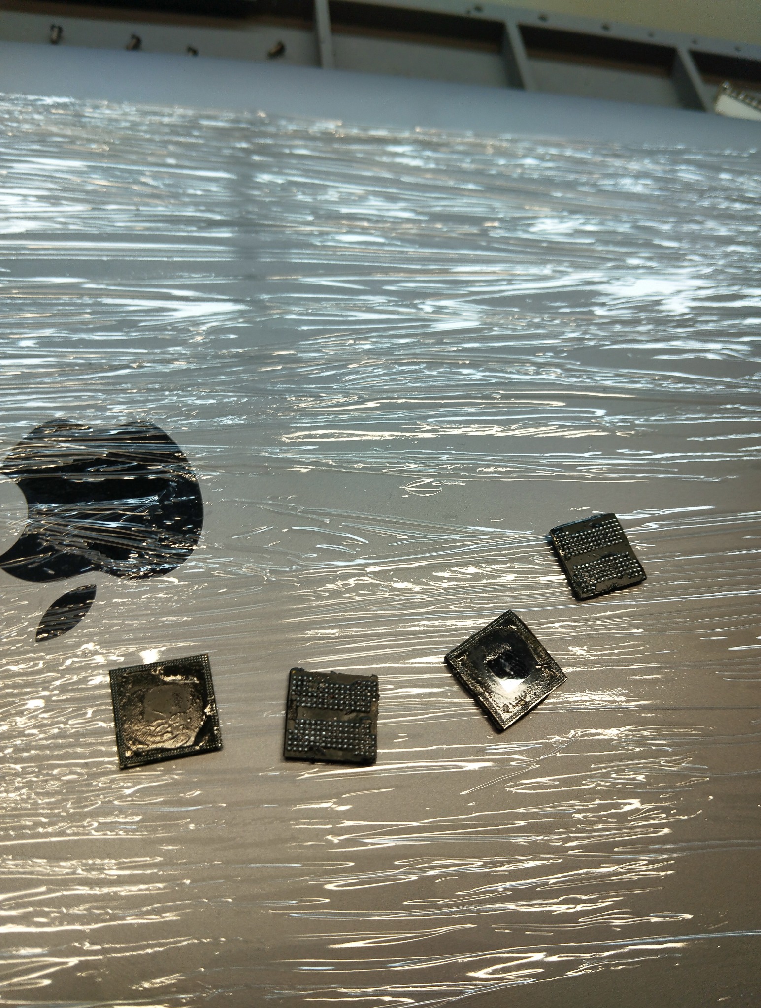 Uszkodzone układy scalone leżące na obudowie laptopa Apple, zabezpieczonej folią. Widoczne ślady uszkodzeń na chipach. Kontekst naprawy elektroniki.