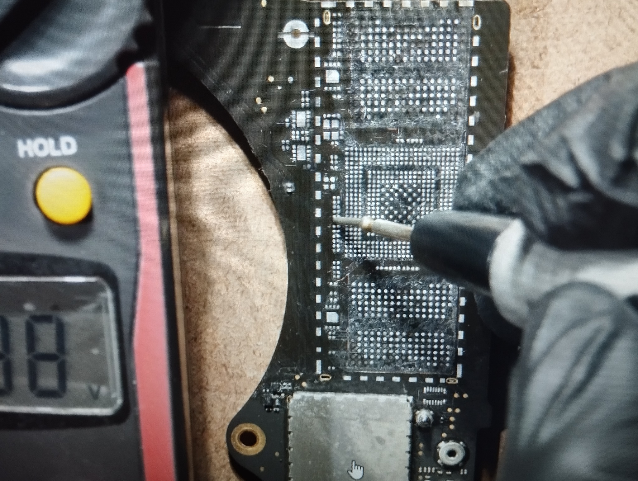 Naprawa płyty głównej: precyzyjne lutowanie układu BGA pod mikroskopem, z użyciem multimetru do pomiaru napięcia. Czarny laminat, białe punkty lutownicze.