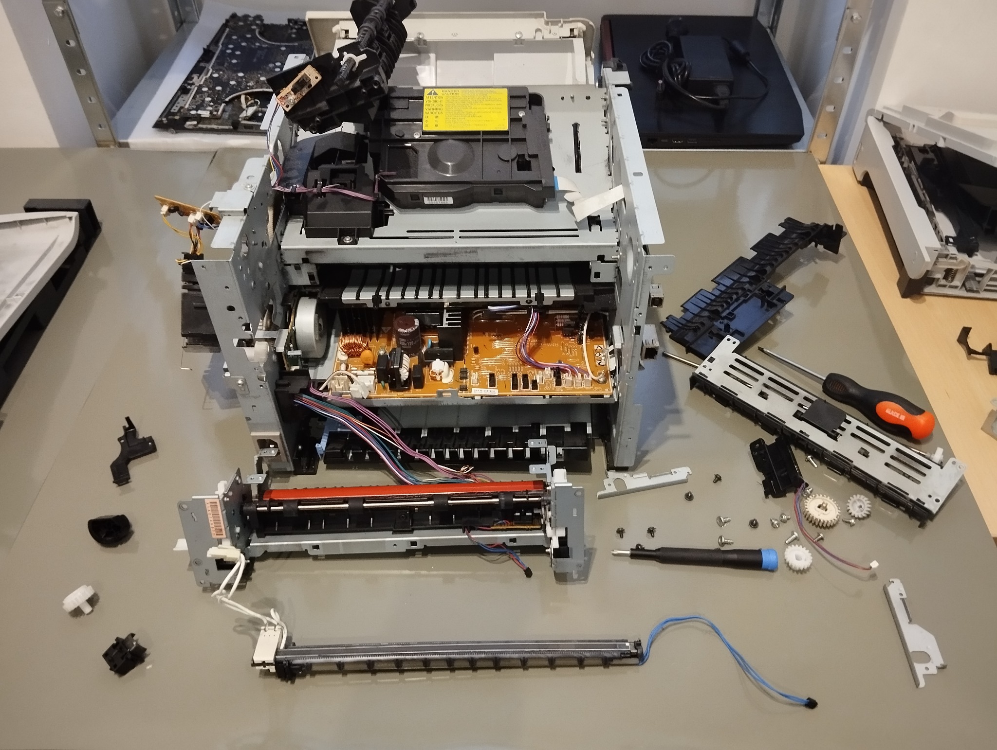 Rozłożona drukarka na stole z narzędziami, ukazująca skomplikowaną konstrukcję i elektronikę podczas naprawy. Widoczne śrubokręty i elementy wewnętrzne.